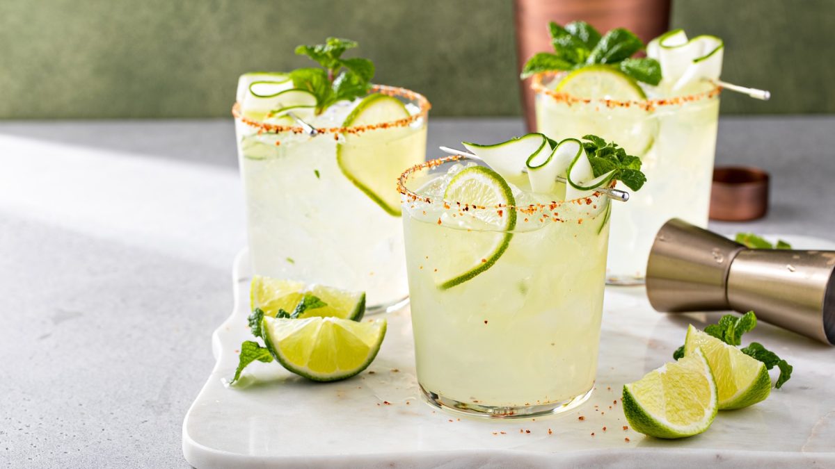 Drei Gläser mit Gurken-margaritas mit Tajin-eand auf einer weißen Marmorplatte, danebebn frische Limettenschnitze, Minze und ein Jigger.