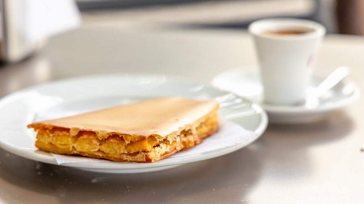 Ein weißer Teller mit dem portugiesischen Gebäck und einer Tasse Kaffee im Hintergrund. Alles auf einem hellen Tisch von der Seite fotografiert.