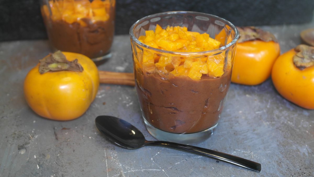 Zwei Gläser mit dem Dessert auf einem Steintisch mit weiteren Kakis, einem schwarzen Löffel und dunklem Hintergrund.