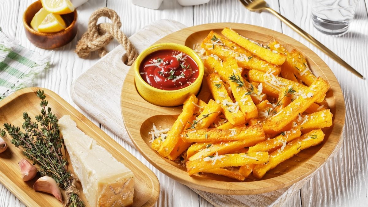 Ein Holzteller mit den Pommes auf einem hellen Tisch mit Kräutern, Parmesan, Gabel, Tuch und weiterer Dekoration. Alles von schräg oben fotografiert.