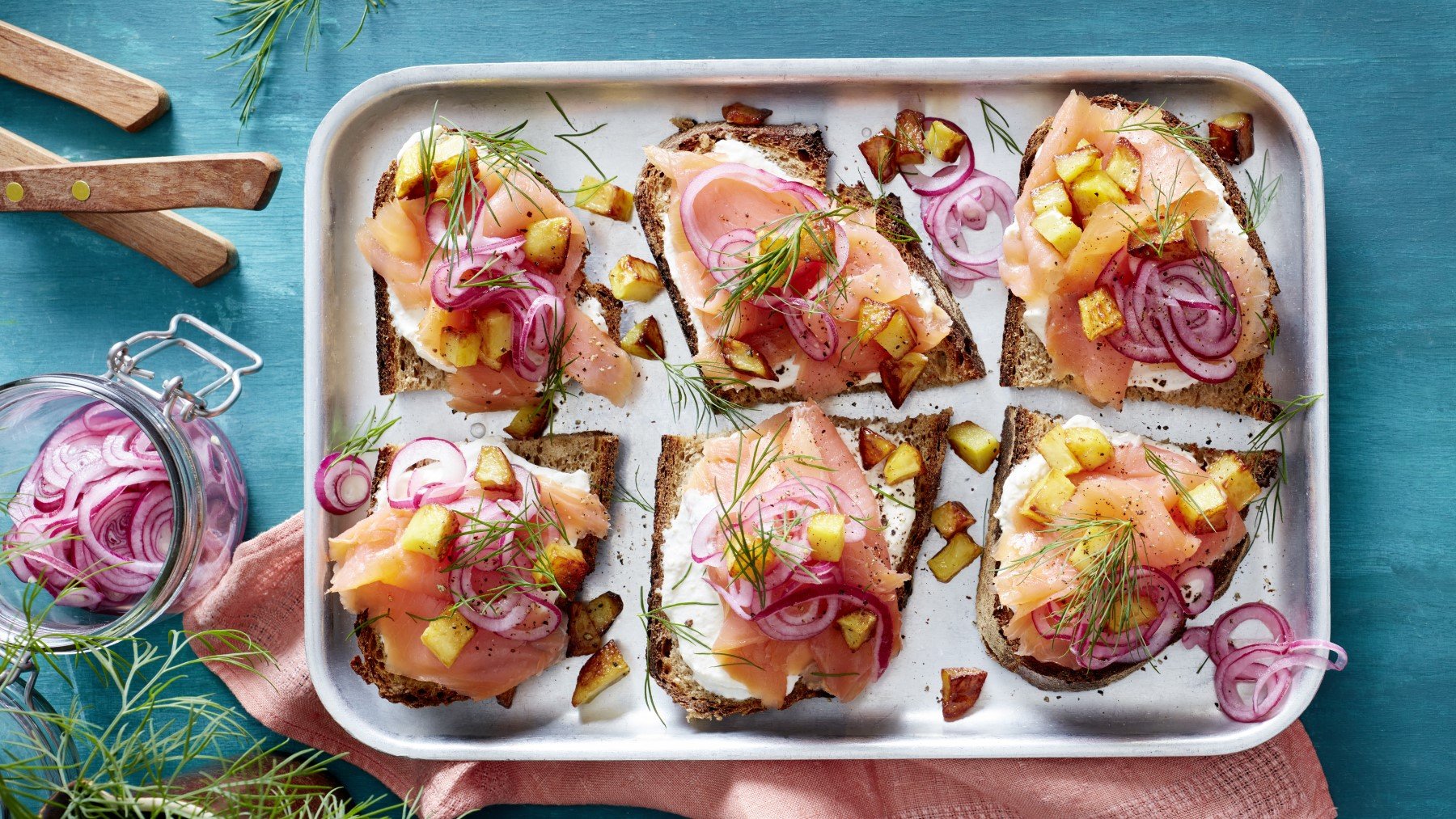 Lachs-Smörrebröd auf einem silbernen Teller auf blauem Hintergrund. Links ein Schälchen mit Zwiebeln, Messer und Dill.