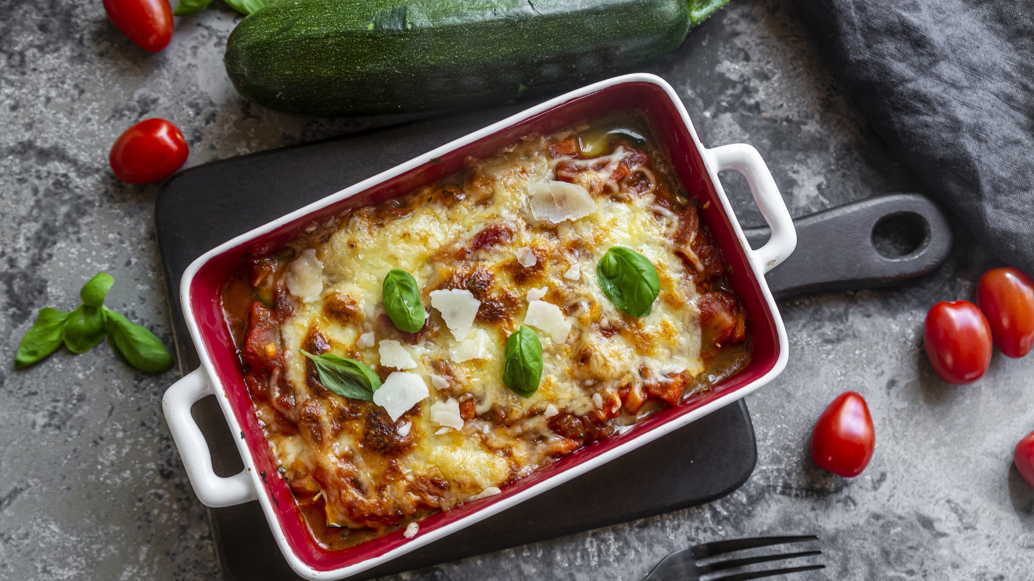 Draufsicht: eine vegetarische Lasagne mit Zucchini in einer weißen Form au einem grauen Untergrund. Drum herum um die Low-Carb-Lasagne liegen frische Basilikumblätter, eine große Zucchini und kleine Tomaten.