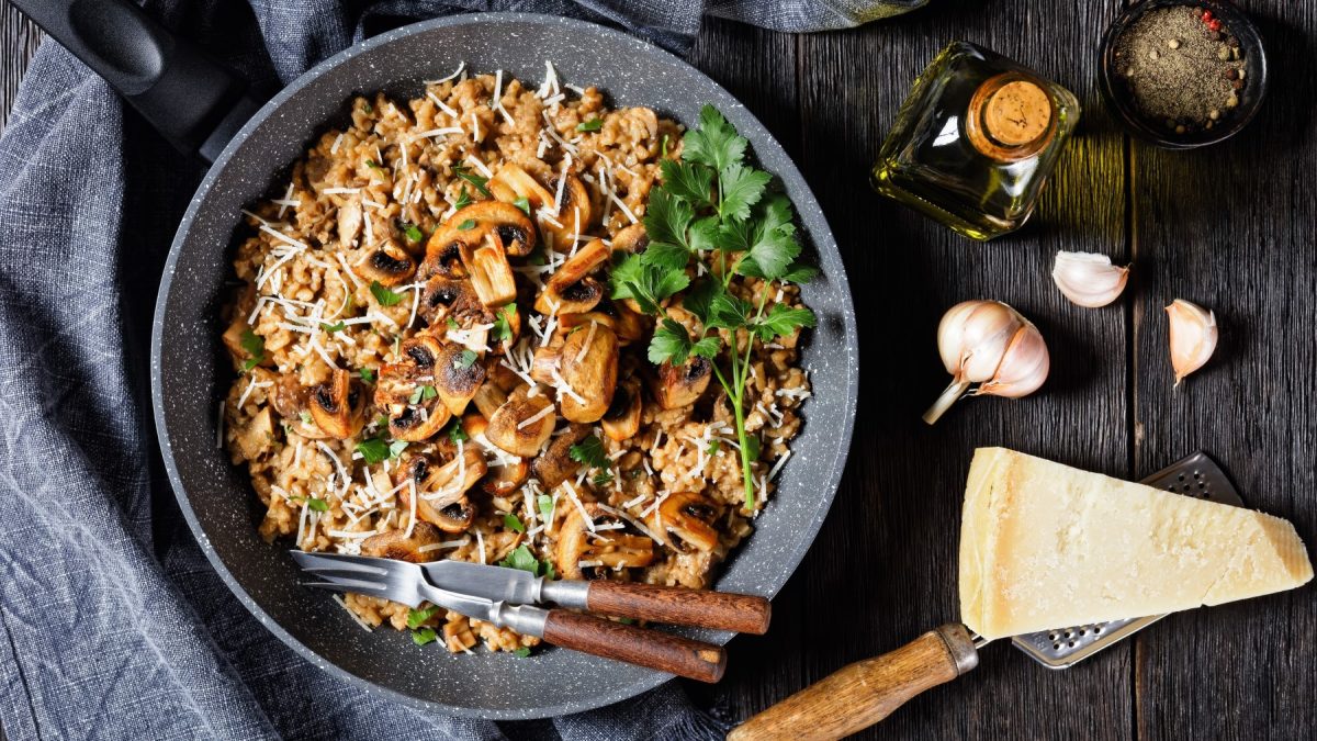 Miso-Pilzrisotto in einem grauen Teller auf dunklem Hintergrund. Links ein graues Geschirrtuch, rechts Knoblauch, Parmesan, Öl und Pfeffer.