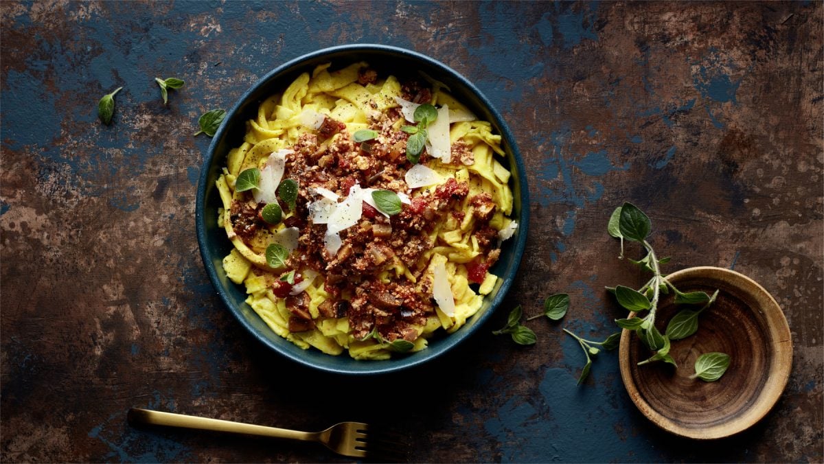 Draufsicht: Schale mit Pappardelle alla Auberginen-Bolo, daneben eine Schale mit frischem Oregano auf dunklem Untergrund.