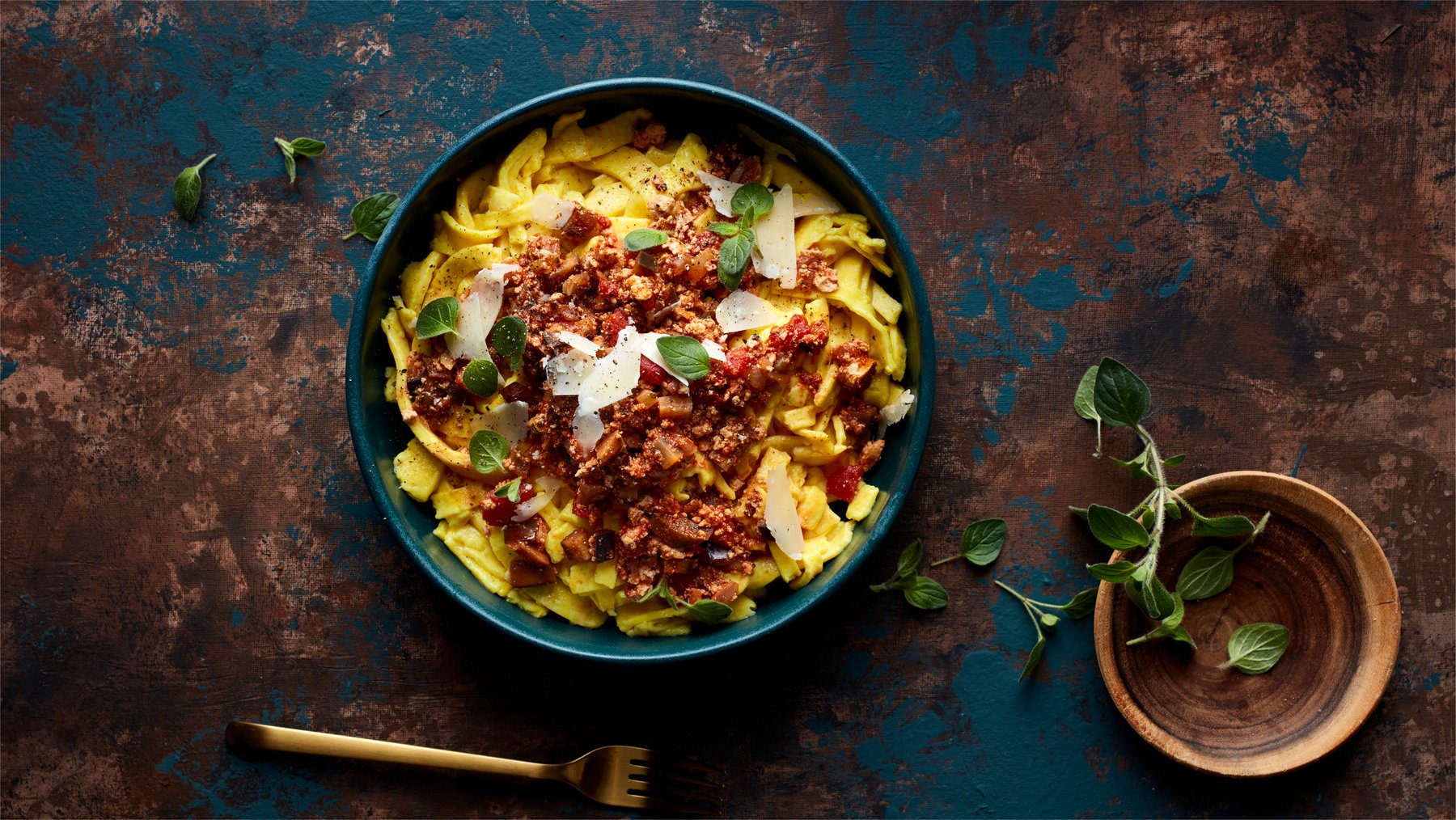 Draufsicht: Schale mit Pappardelle alla Auberginen-Bolo, daneben eine Schale mit frischem Oregano auf dunklem Untergrund.