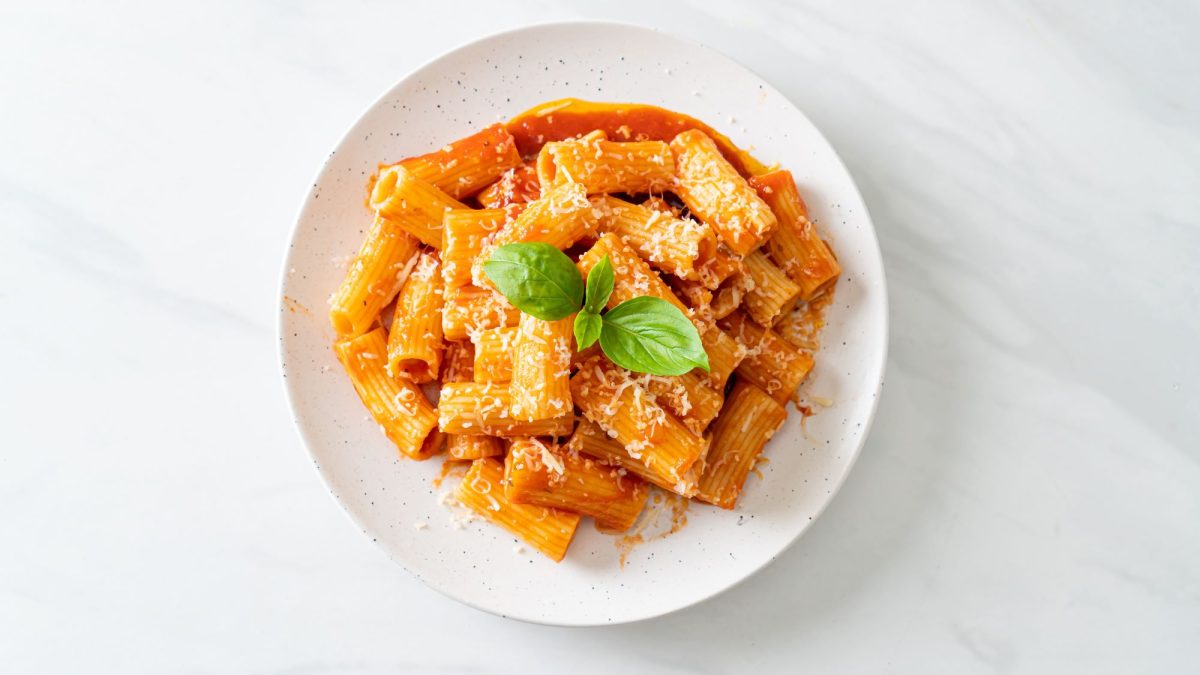 Rigatoni in leuchtend roter Paprikacreme aus dem Ofen mit geriebenem Parmesan und frischem Basilikum, serviert auf einem weißen Teller.