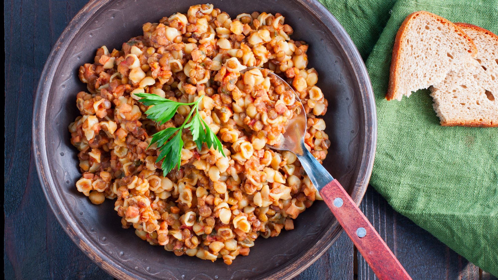 Ein brauner Teller mit kleinen Nudeln in einer würzigen Linsen-Tomatensauce "Pasta e lenticchie", garniert mit frischer Petersilie, auf dunklem Holzuntergrund.