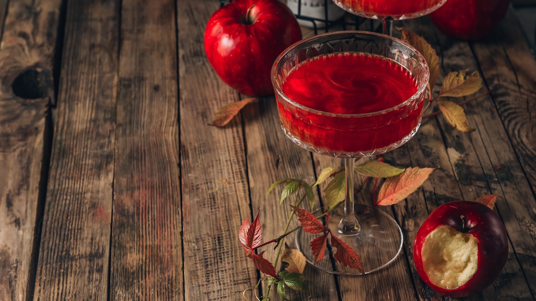 Ein kristallklares Glas mit Poison Apple Cocktail, leuchtende Äpfel und farbige Herbstblätter auf einem rustikalen Holztisch.