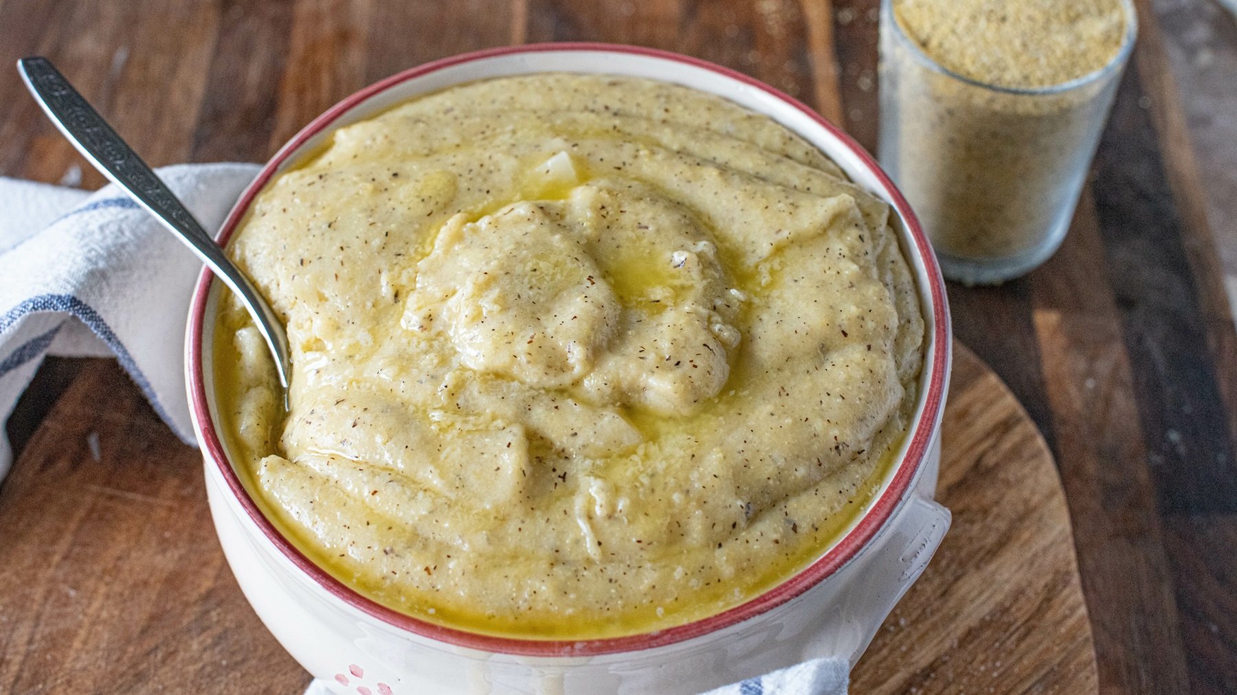 Eine Schüssel mit cremiger Polenta taragna, die mit geschmolzener Butter beträufelt ist, steht auf einem Holzbrett neben einem Glas Polentagrieß.