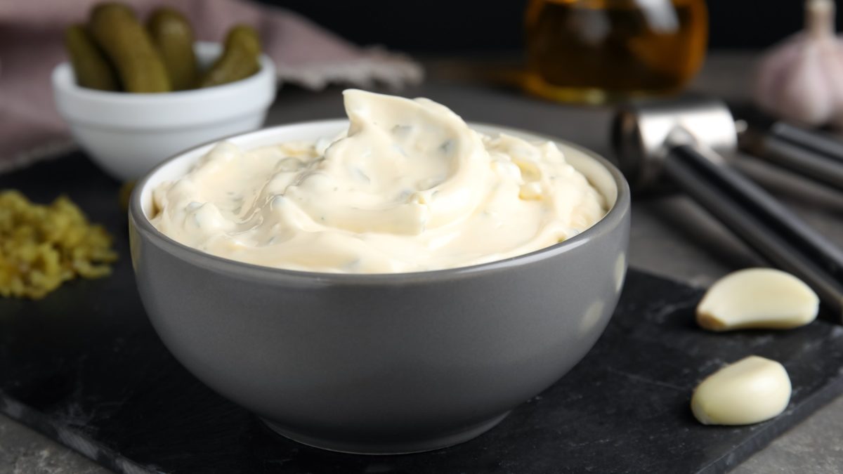 Ein graues Schälchen mit der Sauce Tartare auf schwarzer Schieferplatte und grauem Tisch, mit Zutaten wie Knoblauch, Gewürzgurke und Essig. Alles nah und von der Seite fotografiert.