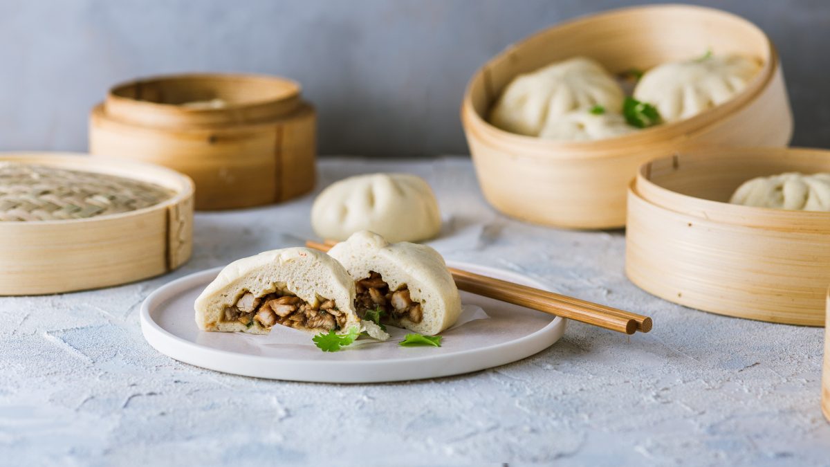 Die Dumplings auf einem hellen Teller und grauem Unter- und Hintergrund mit Dampfkörben und Essstäbchen. Alles von vorne fotografiert.