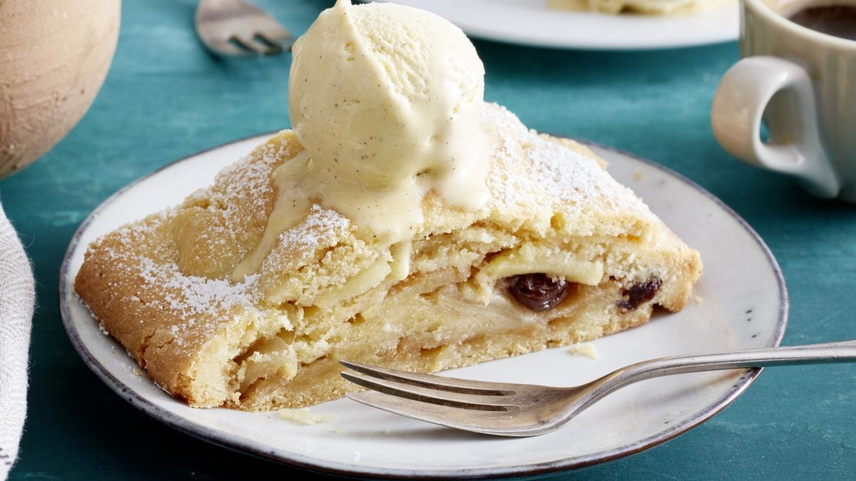 Ein Stück Südtiroler Apfelstrudel auf einem Teller mit Gabel und einer Kugel Vanilleeis oben drauf.