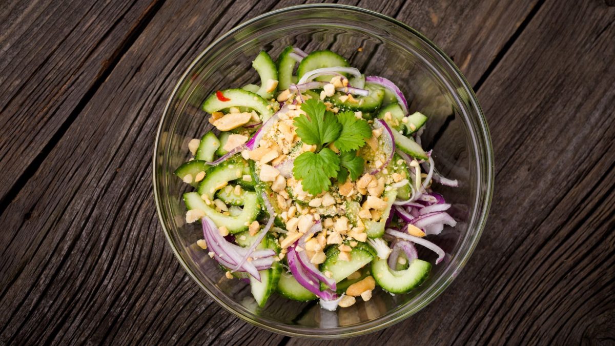 Frischer Thai-Gurkensalat mit roten Zwiebeln, gehackten Erdnüssen und Koriander in einer Glasschale auf rustikalem Holztisch.