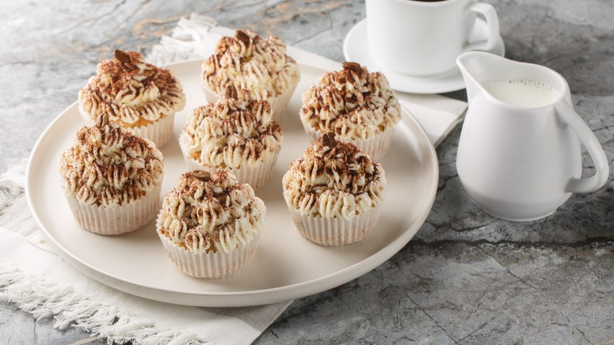 Sieben Tiramisu-Cupcakes auf einem weißen teller, daneben eine Kaffeetasse sowie ein weißes Milchkännchen.