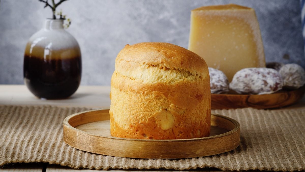 Goldbraunes, luftig gebackene Torta di Pasqua auf einem runden Holztablett, im Hintergrund ein Käseblock, Wurstscheiben und eine Vase.