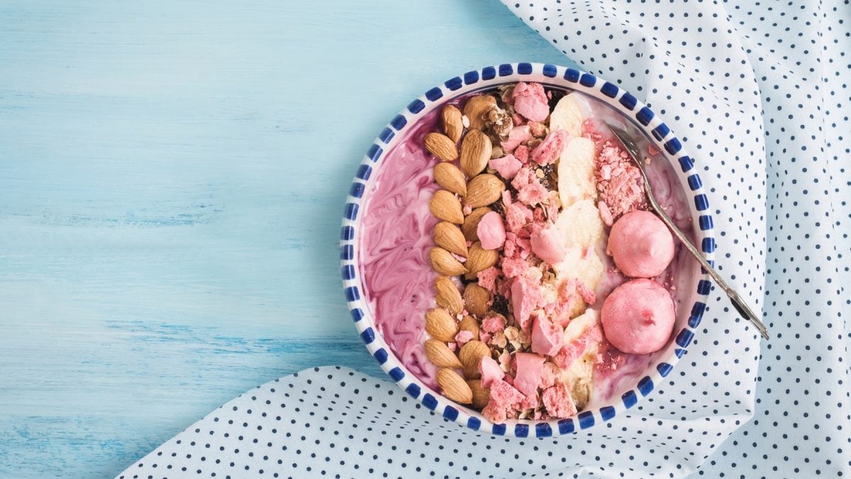 Schale mit Beeren-Joghurtbowl, Mandeln, Bananenscheiben, pinken Fruchtdrops von Sunday Natural und Pralinen auf hellblauem Holztisch mit gepunktetem Tuch.