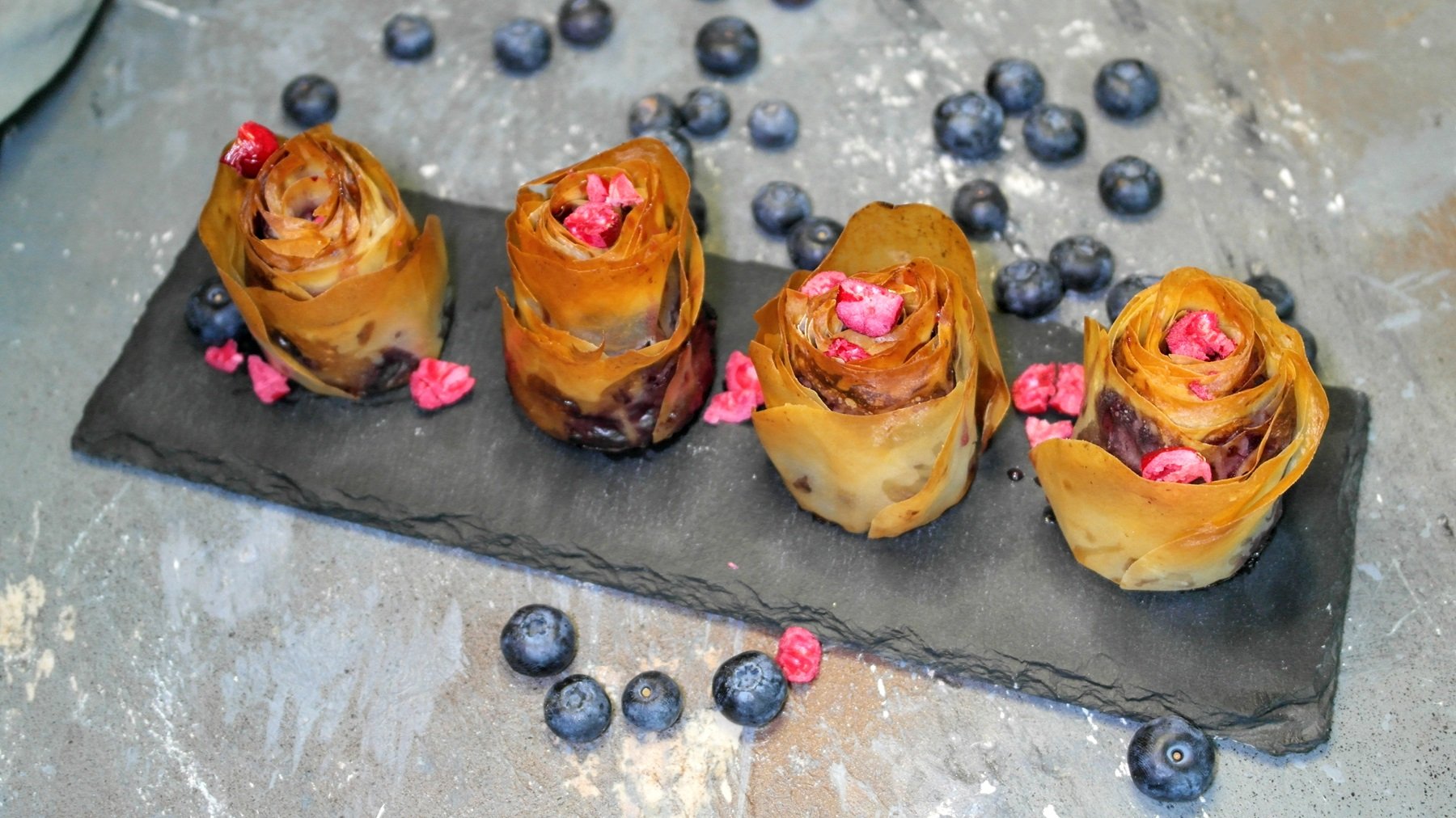 Vier Blaubeer-Rosen auf einer schwarzen länglichen Schiefertafel und grauem Steintisch mit Blaubeeren und grauer Decke. Alles von schräg oben fotografiert.