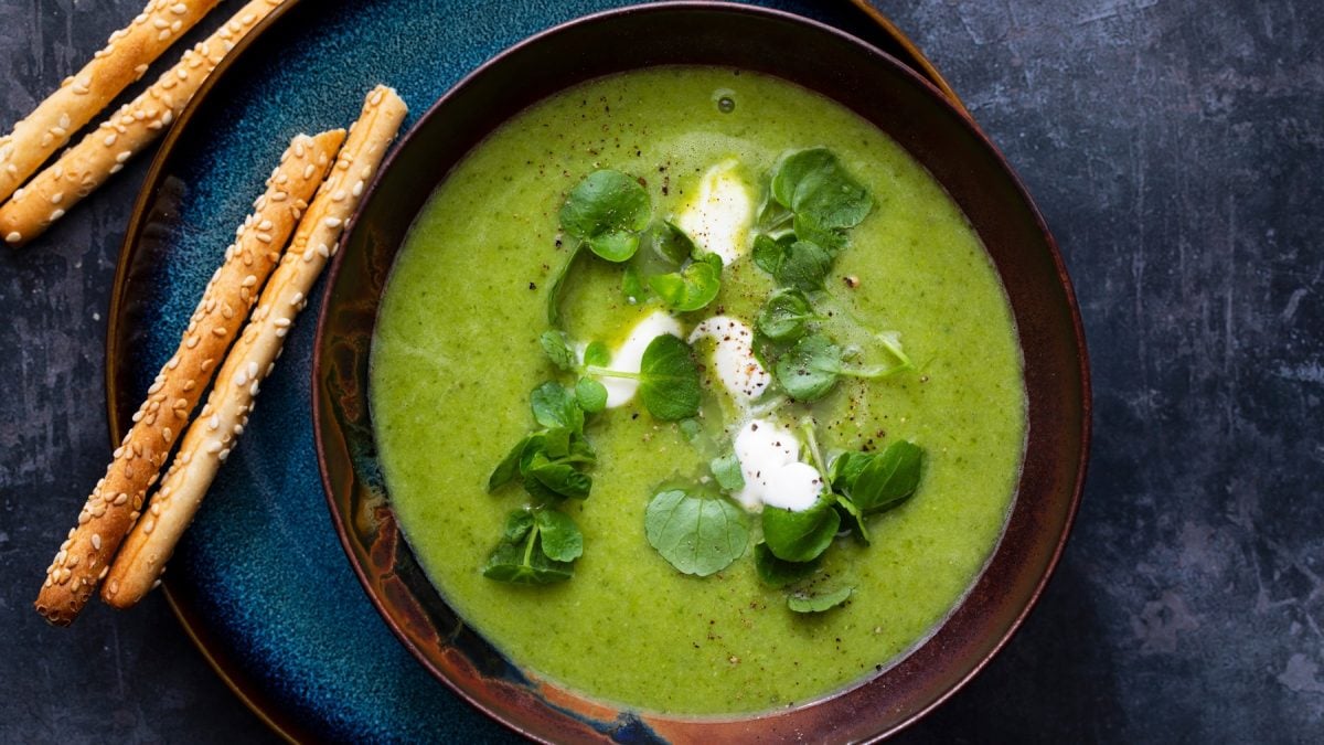 Eine dunkelblaue Schale mit der Brunnenkresse-Suppe auf einem dunklen Tisch. Daneben noch ein paar Gebäckstangen. Alles direkt von oben fotografiert.