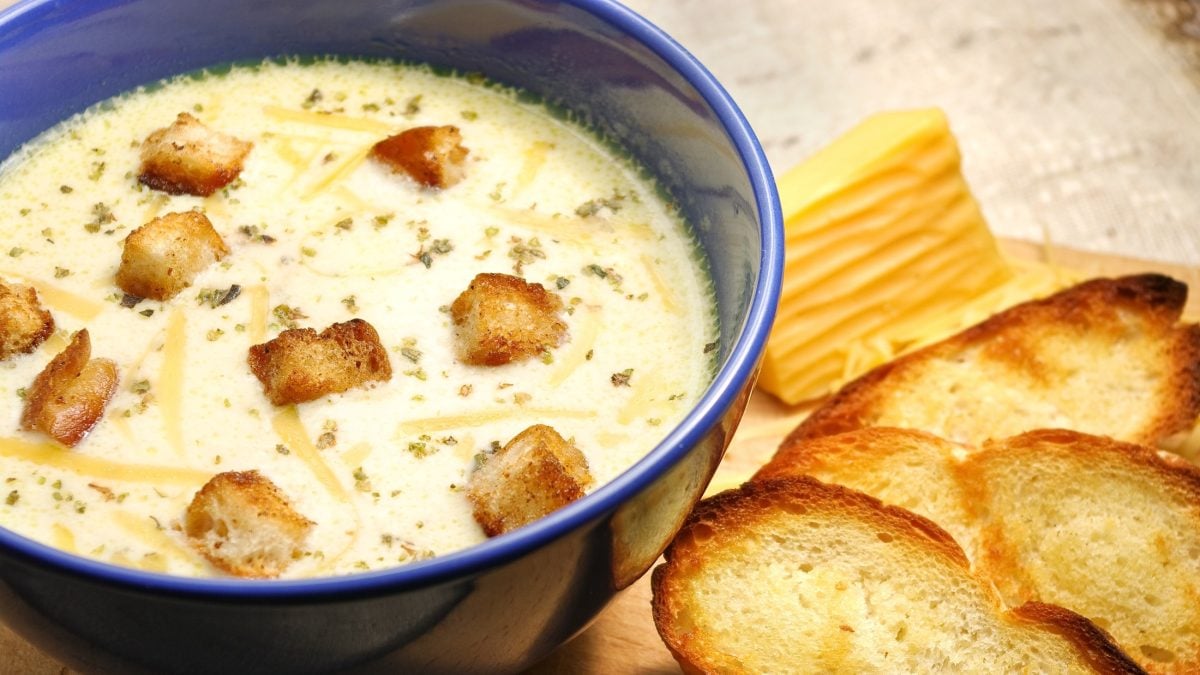 Draufsicht: Eine Käsesuppe in einer runden, dunklen Suppenschüssel. Daneben liegen geröstete Brotscheiben.