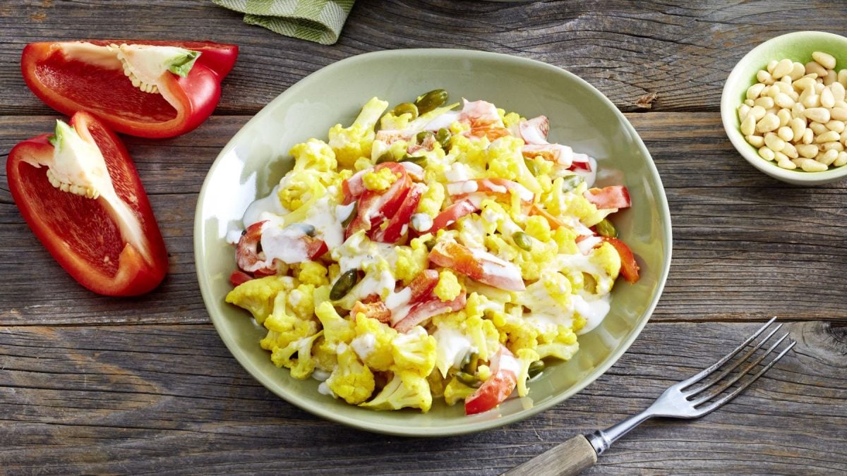 Blumenkohl-Paprika-Salat in einer grünen Schüssel auf hölzernem Hintergrund. Links Paprika und rechts auf dem Foto Pinienkerne.