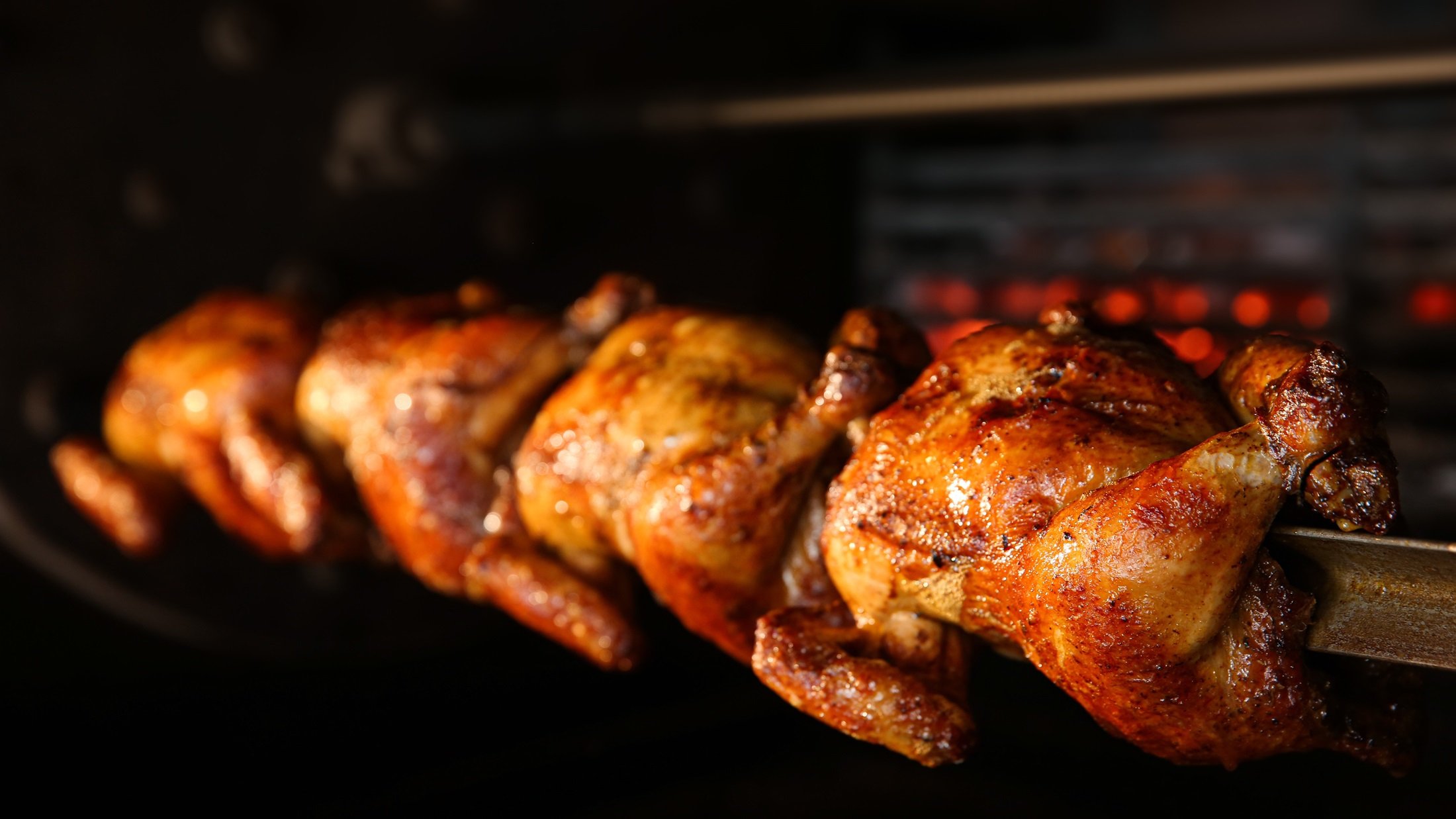 Frontal: Vier Grillhähnchen auf einem Spieß in einem Ofen.