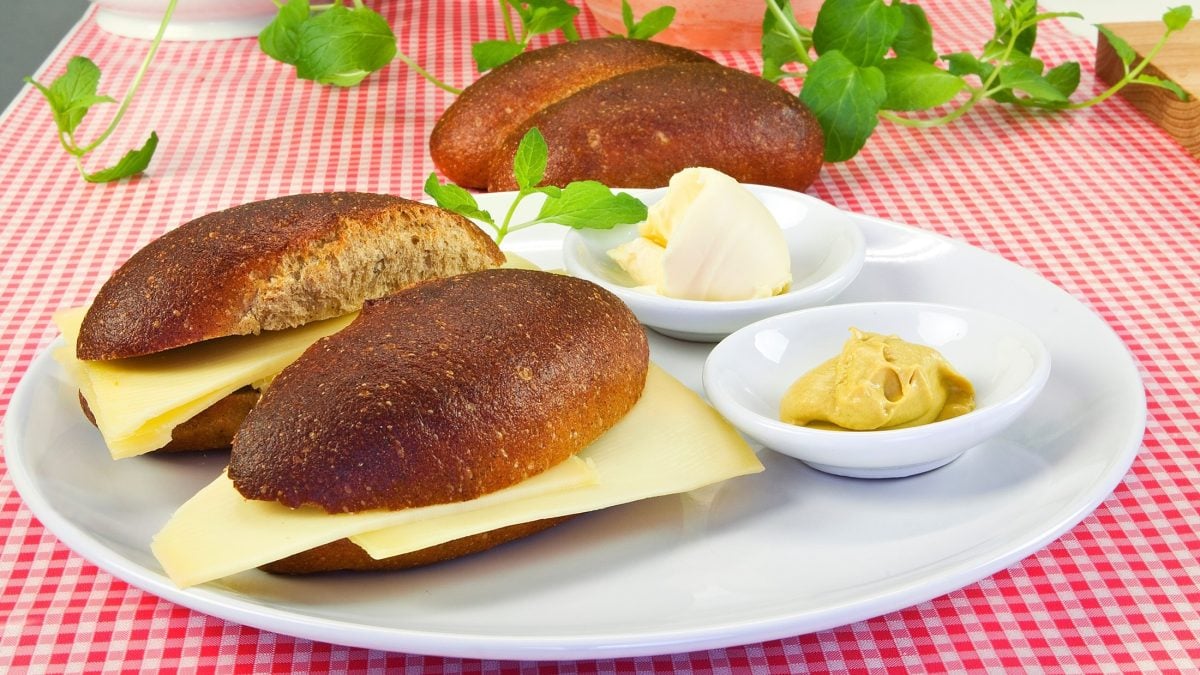 Zwei Käsebrötchen auf einem hellen Teller. Im Hintergrund ist ein Brötchen ohne Käse. Auf dem Teller ist eine kleine Schale mit Senf und eine Mit Butter.