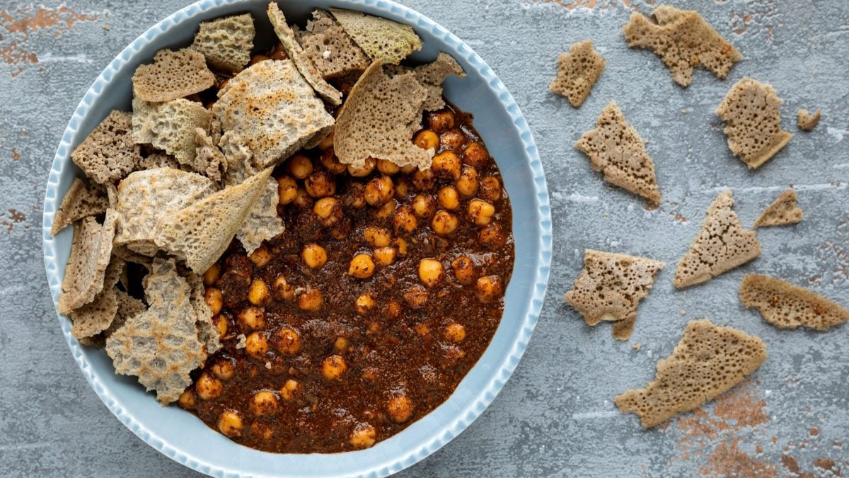 Draufsicht: Auf hellem Grund aus Holz steht eine runde Schale mit gekochten Kichererbsen und kleinen gezupften Brotstücken, Weitere kleine Brotfetzen liegen rechts neben der Schüssel.