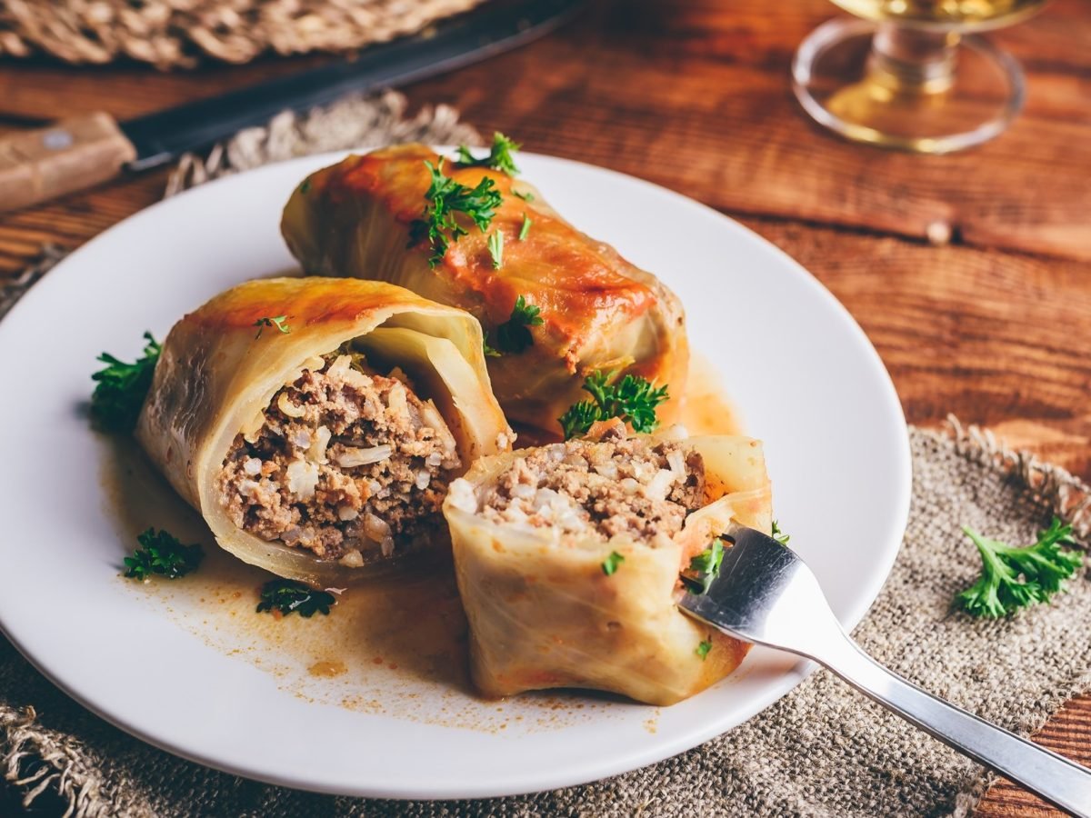 leicht schräge Draufsicht: Zwei Kohlrouladen mit Hackfleisch-Füllung auf einem hellen runden Teller. Eine Roulade ist halbiert. In einer Hälfte steckt eine Gabel. Der Untergrund ist aus Holz.