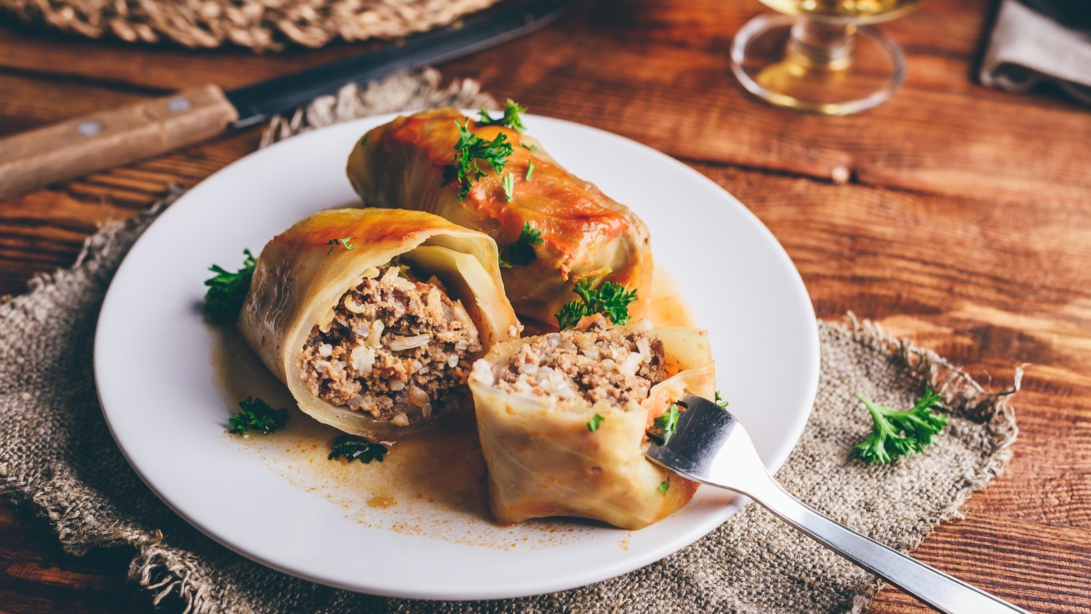 leicht schräge Draufsicht: Zwei Kohlrouladen mit Hackfleisch-Füllung auf einem hellen runden Teller. Eine Roulade ist halbiert. In einer Hälfte steckt eine Gabel. Der Untergrund ist aus Holz.