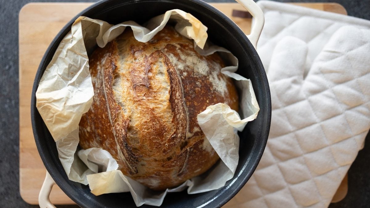 Draufsicht: Ein rundes gebackenes Brot in einem runden, offenen Topf. Der steht auf einem Holzbrett. Daneben liegt ein Topflappen.