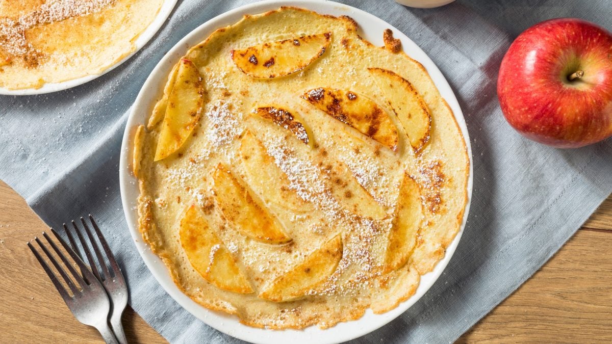 Draufsicht: Ein runder Pfannkuchen mit Apfel auf einem runden Teller. Neben dem liegt ein frischer Apfel.