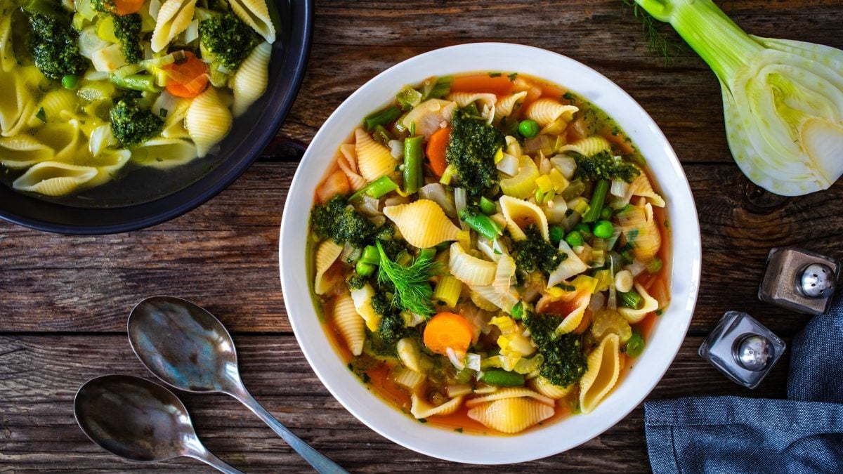 Draufsicht: Auf einem dunklen Untergrund aus Holz steht eine runder Eintopf, in dem eine Gemüsesuppe ist. Drum herum liegen Suppenlöffel und frisches Gemüse.