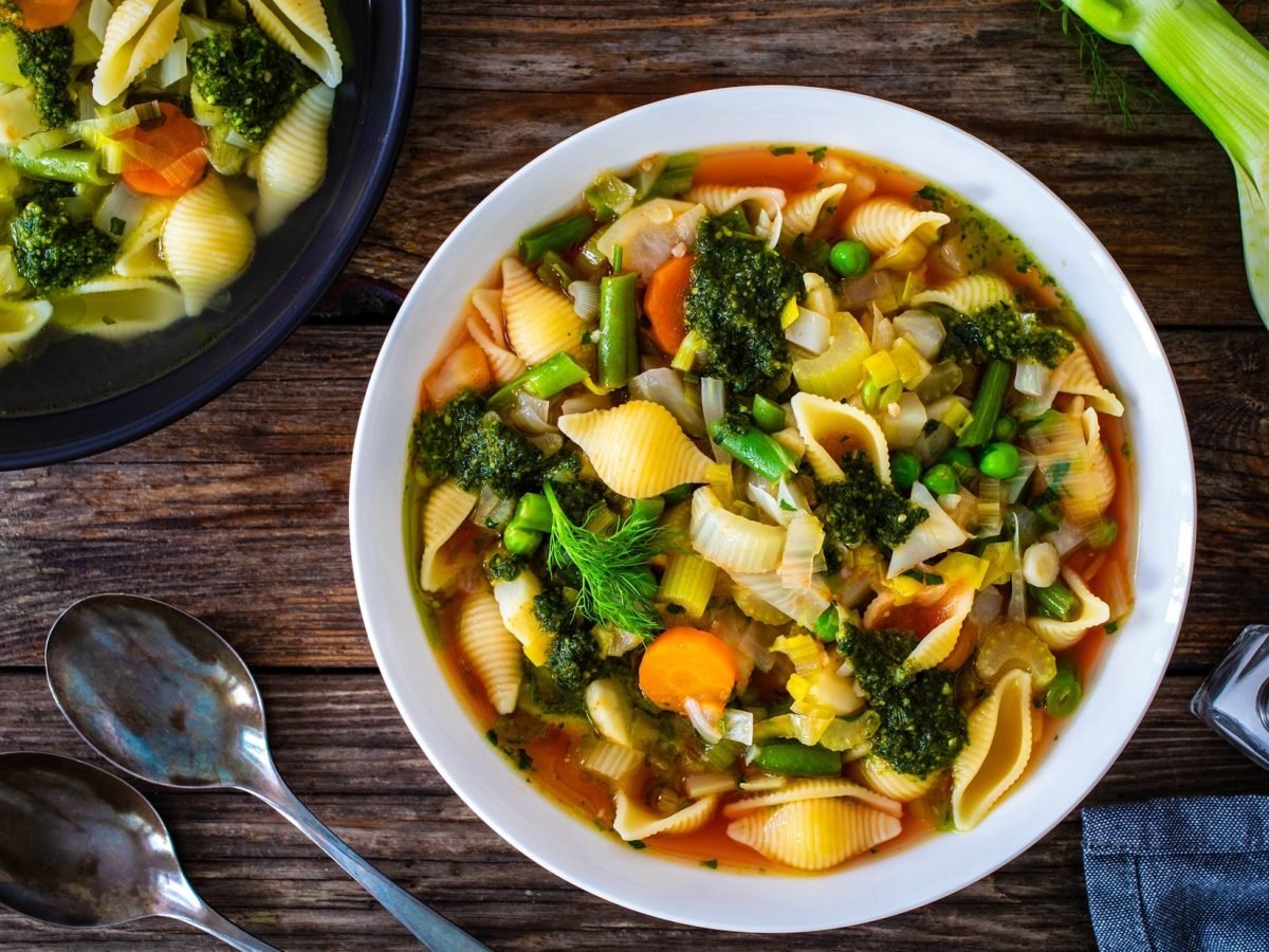 Draufsicht: Auf einem dunklen Untergrund aus Holz steht eine runder Eintopf, in dem eine Gemüsesuppe ist. Drum herum liegen Suppenlöffel und frisches Gemüse.