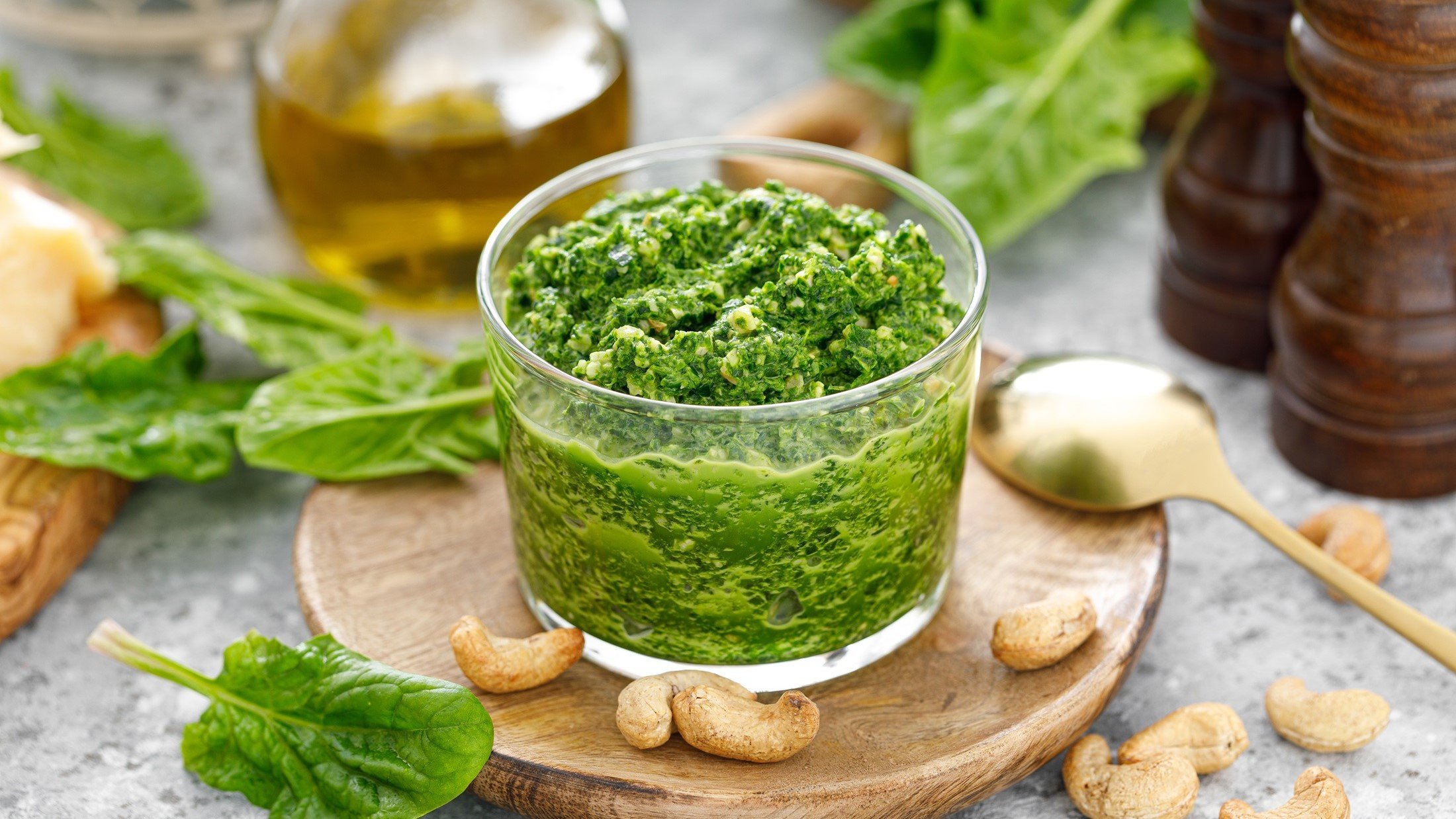 Draufsicht, leicht schräg: Ein offener Behälter aus Glas mit einem Spinat-Pesto mit Cashews. Drum herum liegen oder stehen u.a. frische Spinatblätter, Cashewkerne, eine Karaffe mit Öl, eine Suppenlöffel und zwei Gewürzmühlen aus Holz.