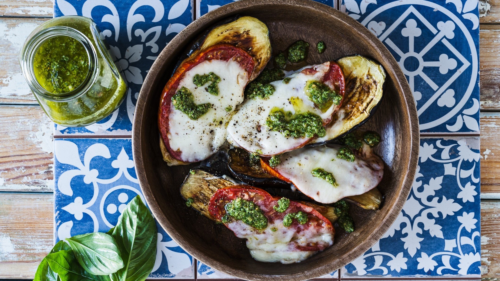 Draufsicht: Ein runder Teller. Auf der liegen vier mit Mozzarella überbackenen Scheiben Aubergine. Daneben liegen frische Basilikumblätter und eine kleines Glas mit einem Kräuteröl, dass an ein Basilikumpesto erinnert.