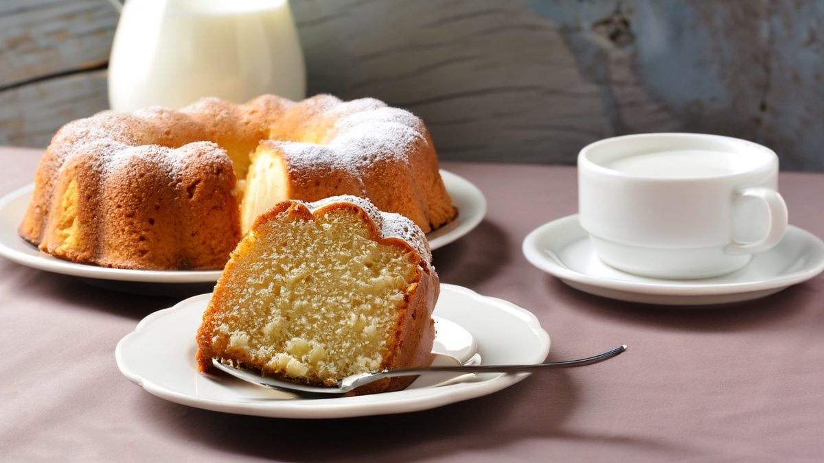 Ein frisch gebackener französischer Joghurtkuchen mit Puderzucker, ein Stück davon auf einem Teller mit Löffel, daneben eine Tasse Milch.