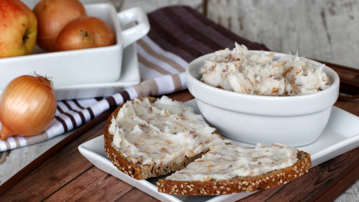 Frontal: Auf einem Holztisch stehen eine helle, runde Schüssel mit Griebenschmalz. Daneben liegen zwei halbe Brotscheiben mit dem Aufstrich. Im Hintergrund sind frische Zwiebeln.