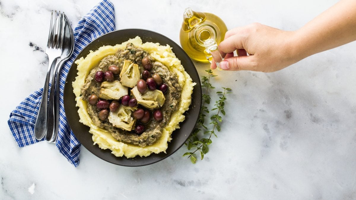 Hand gießt Öl über Kartoffelpüree mit Artischocken, Oliven und Baba Ganoush auf schwarzem Teller, daneben blaue Serviette.