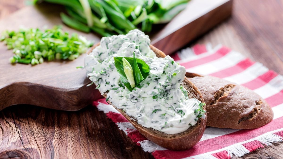 Draufsicht: Auf einem Holztisch liegen ein Brot mit Kräuterquark und ein Küchenhandtuch. Drum herum sind frische Kräuter.