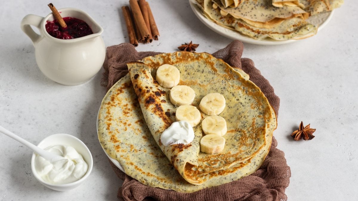 Nahaufnahme eines Mohnpfannkuchens mit karamellisierten Stellen, serviert mit Bananen, Joghurt und Gewürzen auf hellem Steinhintergrund.