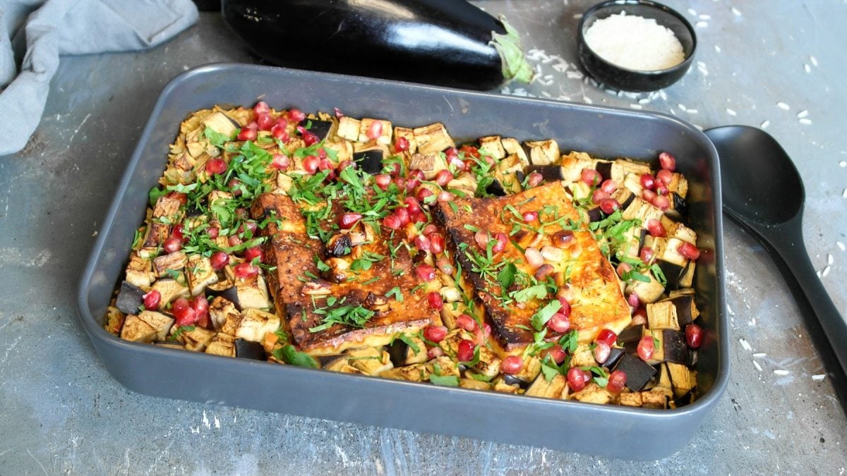 Eine graue Auflaufform mit dem Reisgericht auf einem Steintisch. Daneben ein schwarzer Kochlöffel und dahinter eine Aubergine sowie ein Schälchen mit Reis. Alles von schräg oben fotografiert.