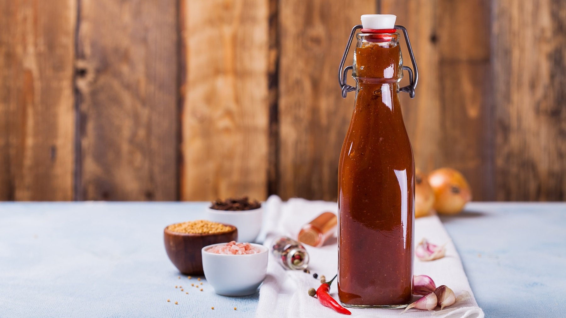 Eine Flasche mit der abgefüllten Schaschlik_Sauce auf einem grauen Tisch und hölzernem Hintergrund. Alles von der Seite fotografiert. Um die Flasche herum liegen noch Kochutensilien und Zutaten.