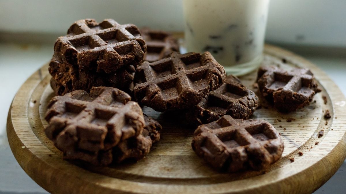 Die Schoko-Waffel-Kekse auf einem runden Schneidebrett mit einem Glas Milch im Hintergrund. Alles auf hellem Untergrund fotografiert.