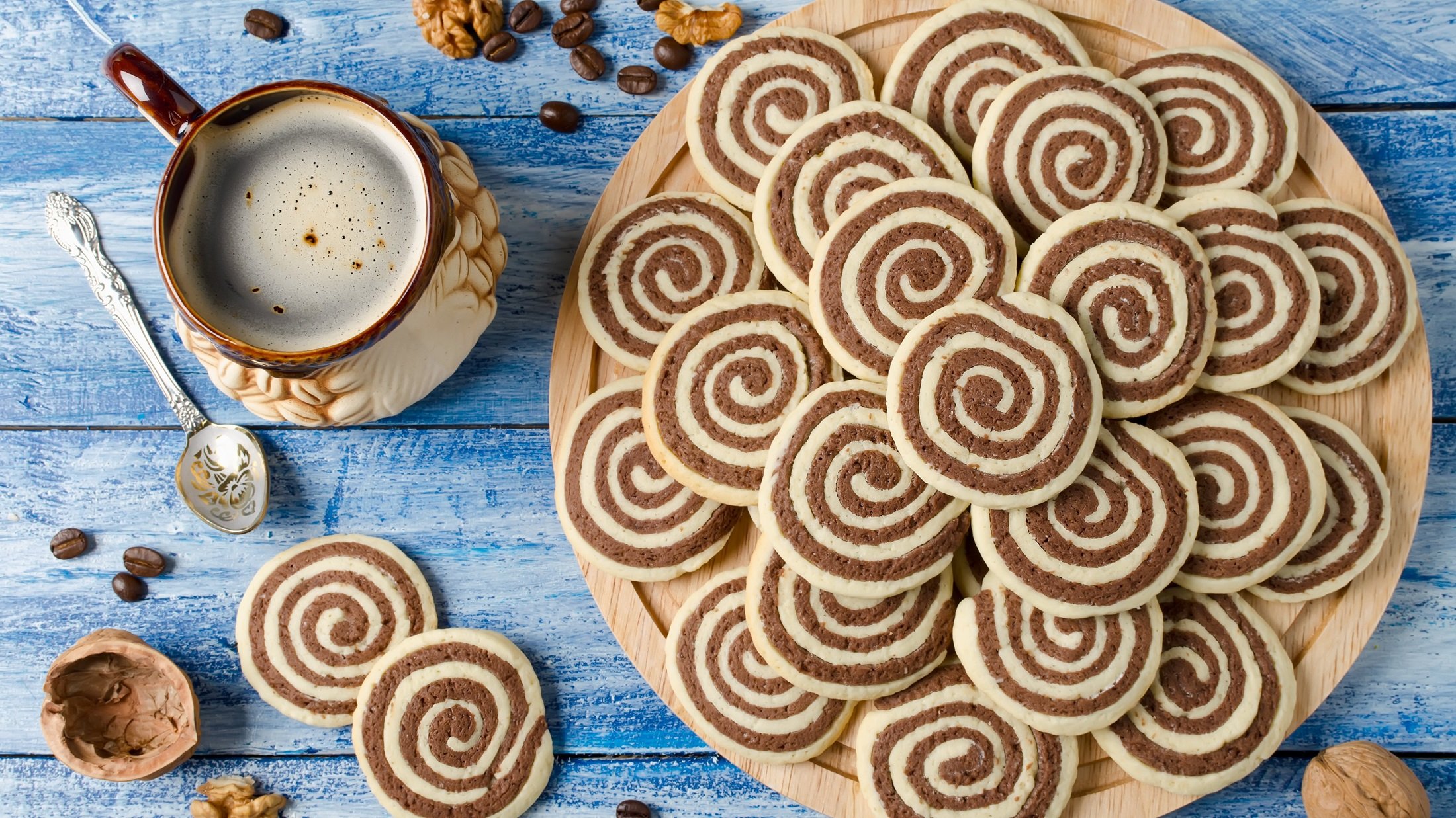 Draufsicht: Auf einem Untergrund aus Holz liegt ein rundes Holzbrett auf dem runde Kekse sind. Daneben liegen weitere Kekse, eine Tasse mit Kaffee, Kaffeebohnen und Nüsse.