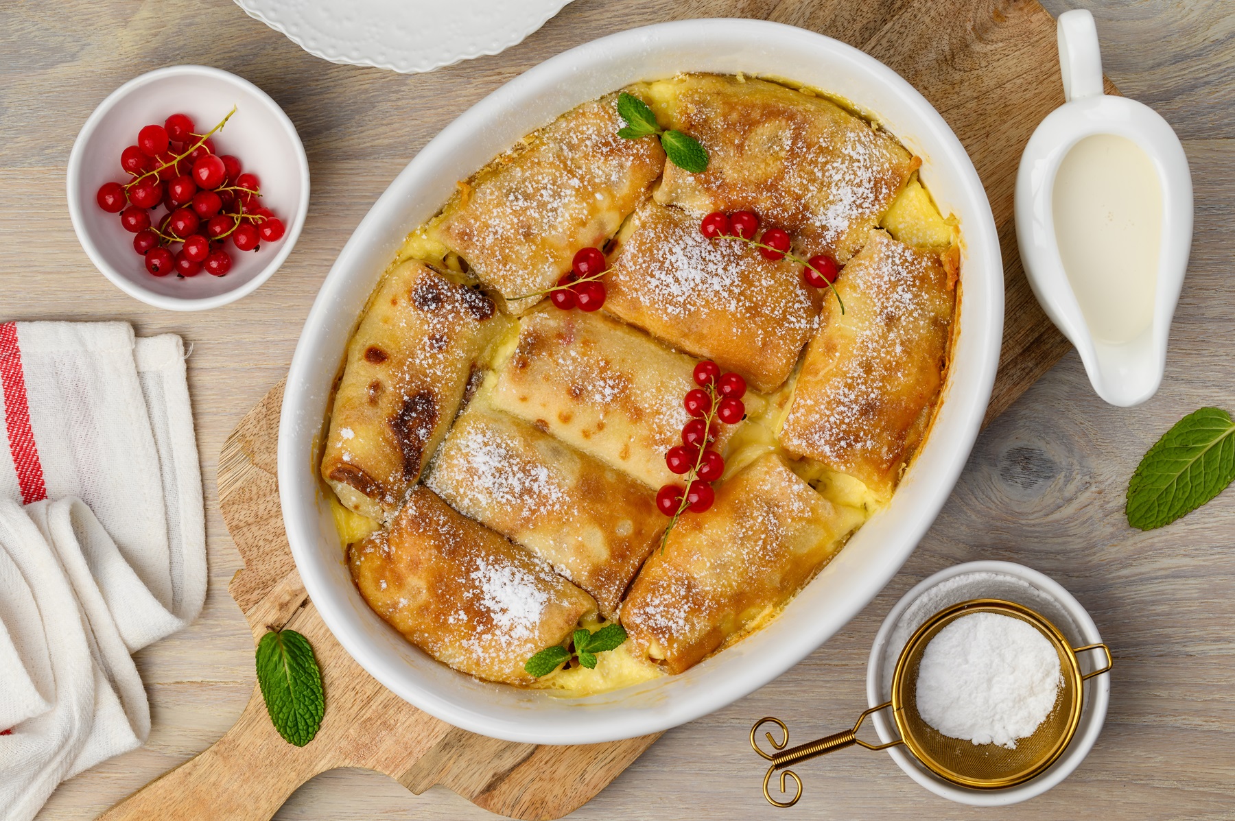 Frisch gebackene goldbraune Topfenpalatschinken in einer weißen Auflaufform, mit Puderzucker bestäubt und mit roten Johannisbeeren garniert.