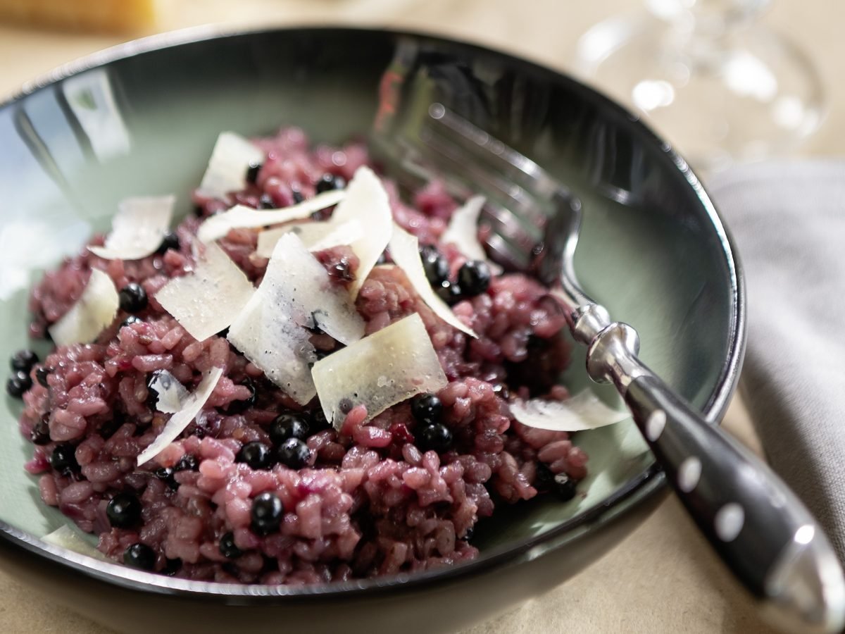 Ein dunkler, tiefer Teller mit dem violetten Holunder-Risotto auf einem beigen Tisch.