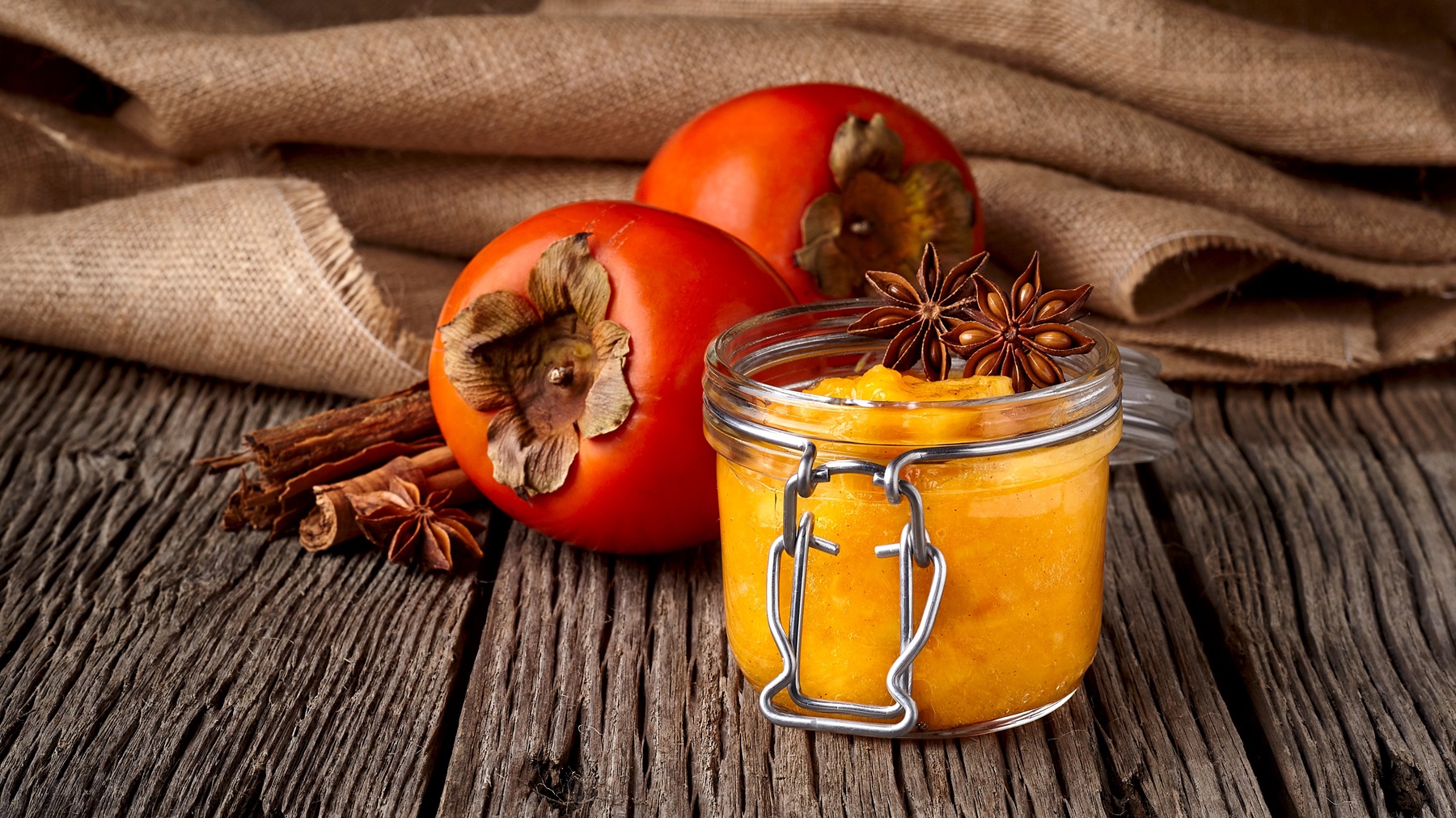 Eimn Glas mit der MArmelade auf einem dunklen Holztisch mit Kaki und Zimtstangen im Hintergrund.